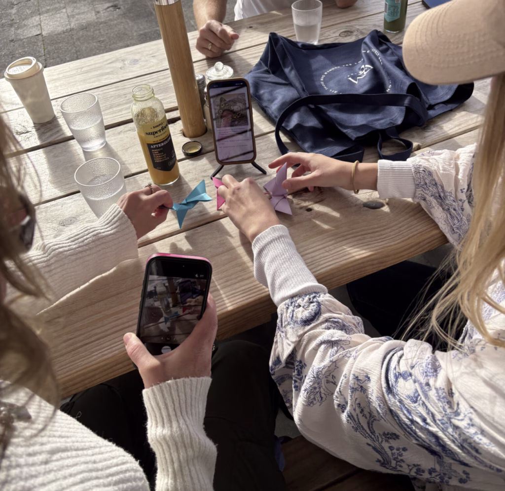 Two women look at a phone screen and create origami shapes with paper. They seem to be sitting at a picnic table with coffee, a smoothie, and waters and one woman has her camera app open.